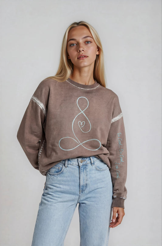 Woman wearing a House-of-Malibu brown taupe sweatshirt with a painted buddha heart design and light blue jeans on a plain background
