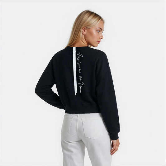 Back view of a woman wearing a Black cozy sweatshirt in a cropped length and a back stripe with a manifesting universe handwritten quote.