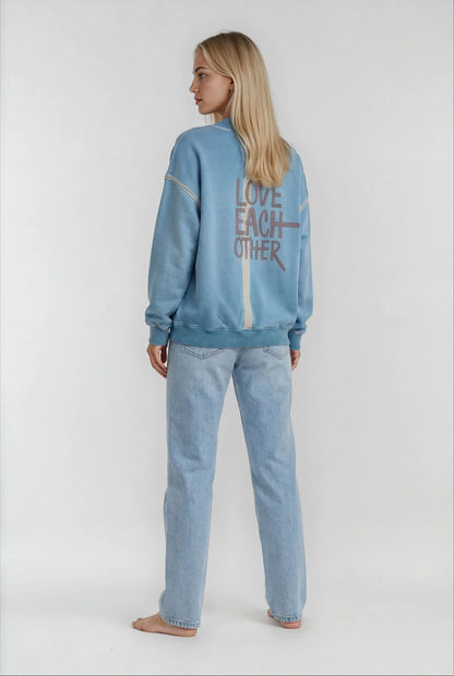 Person wearing a house of malibu light blue sweatshirt with 'LOVE EACH OTHER' text on the back, standing against a white background.