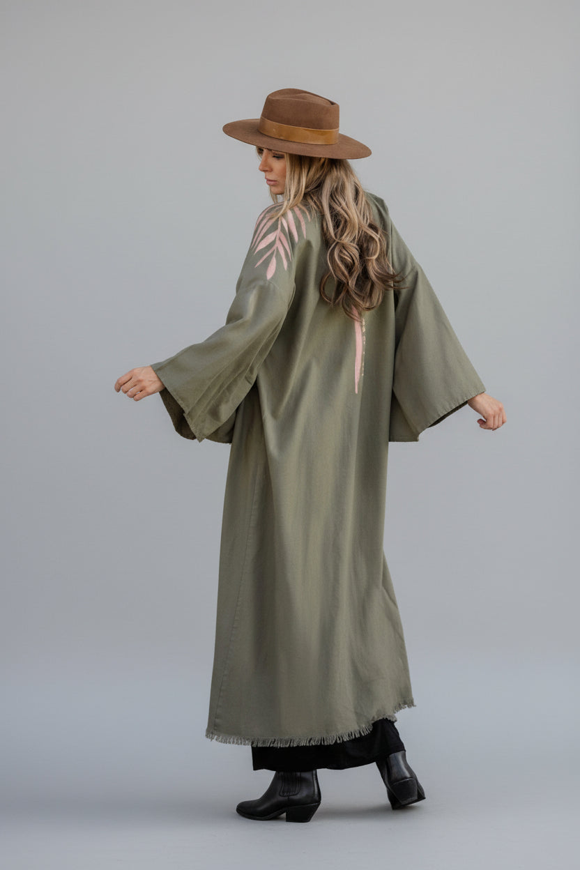 A person wearing an olive green hand-painted linen kimono with a floral pattern and text on the back that says 'Take me as I am'.