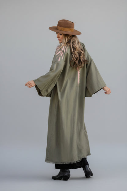 A person wearing an olive green hand-painted linen kimono with a floral pattern and text on the back that says 'Take me as I am'.
