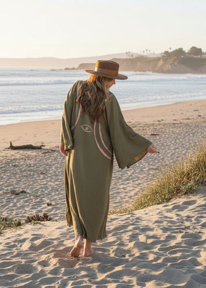 A woman on the beach walking in a long olive kimono hand-painted lines and an eye motif  showing her from behind