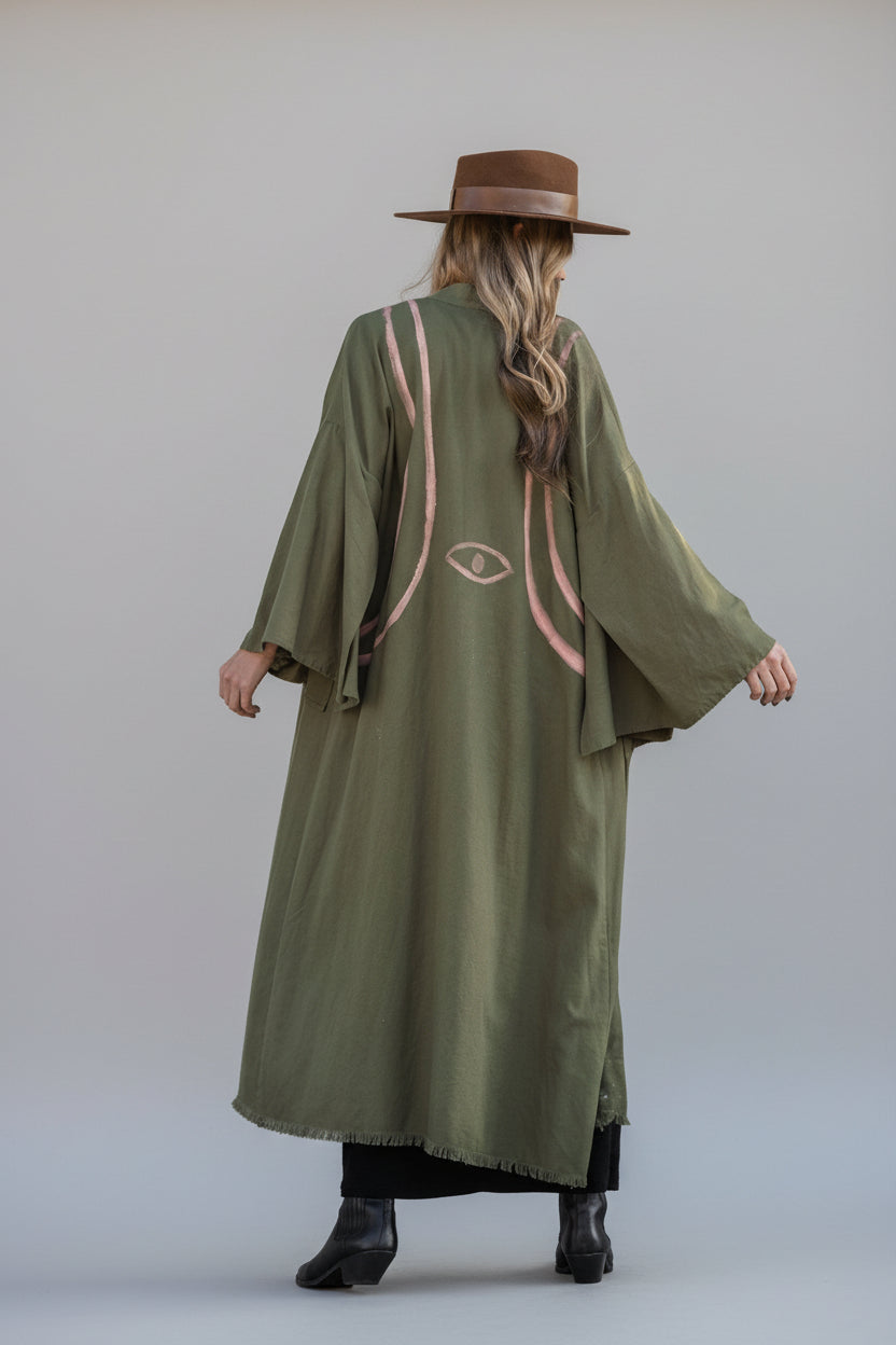 A woman styled in an olive maxi length kimono which has hand-painted blush designs of lines which harness her body from back to front and an evil eye centered on her back
