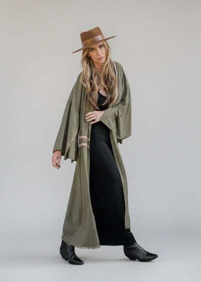 A woman wearing a hand-painted  olive blush hand-painted linen kimono with a wide-brimmed hat, standing against a gray wall.