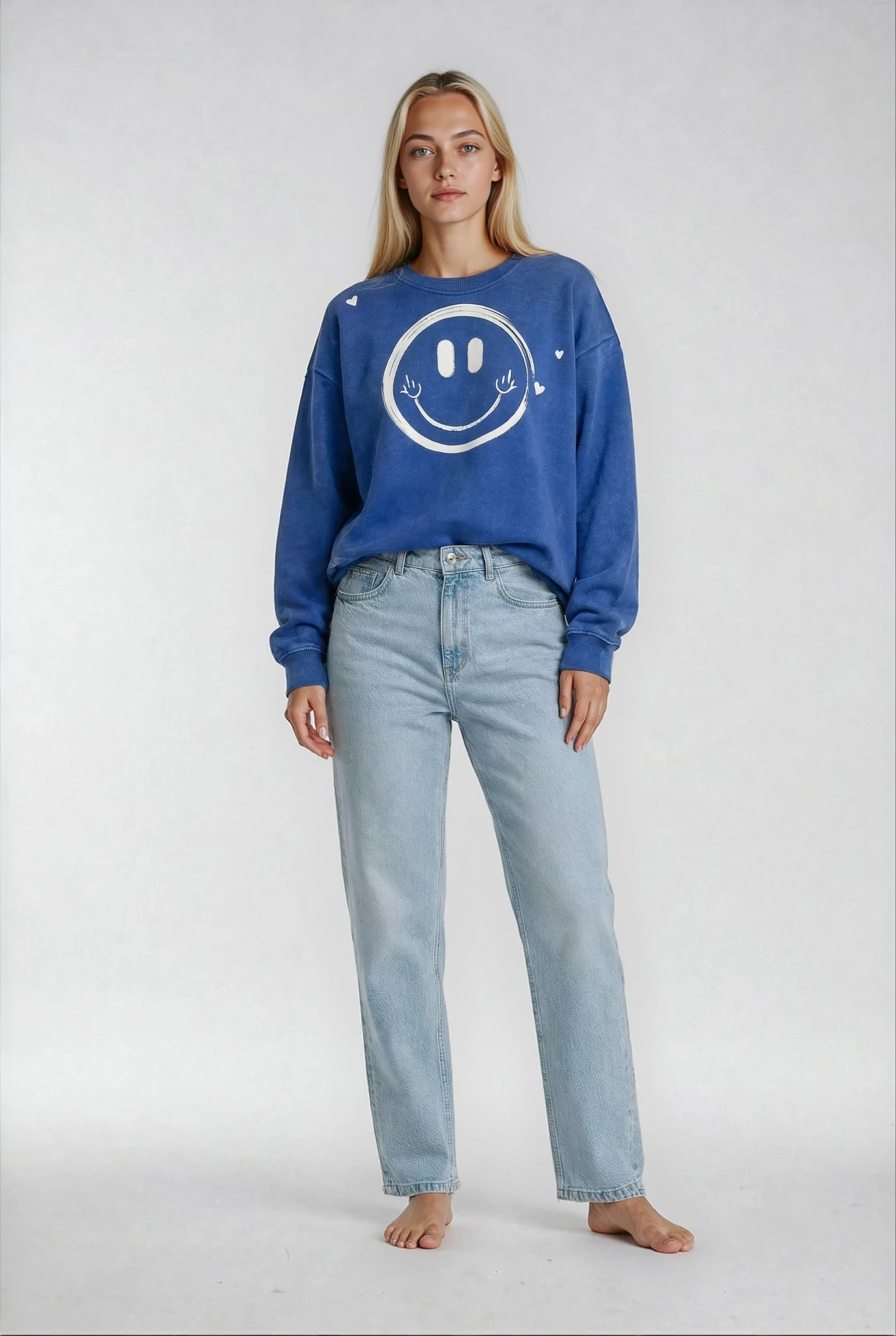 Woman wearing a royal blue sweatshirt with a smiley face design on the front and hearts around it   by brand House of Malibu and light blue jeans