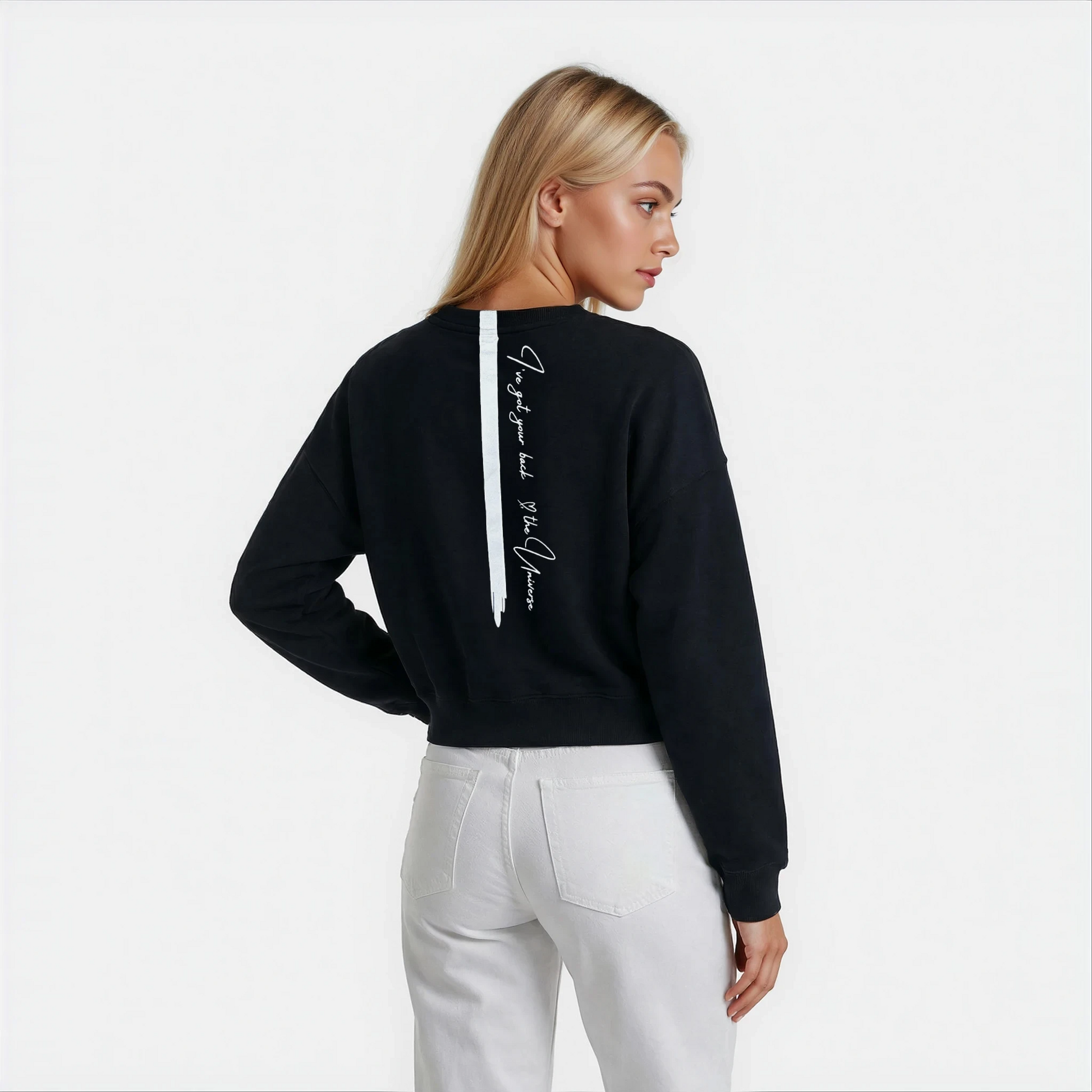 Back view of a woman wearing a Black cozy sweatshirt in  a cropped length and a back stripe with a manifesting universe handwritten quote.