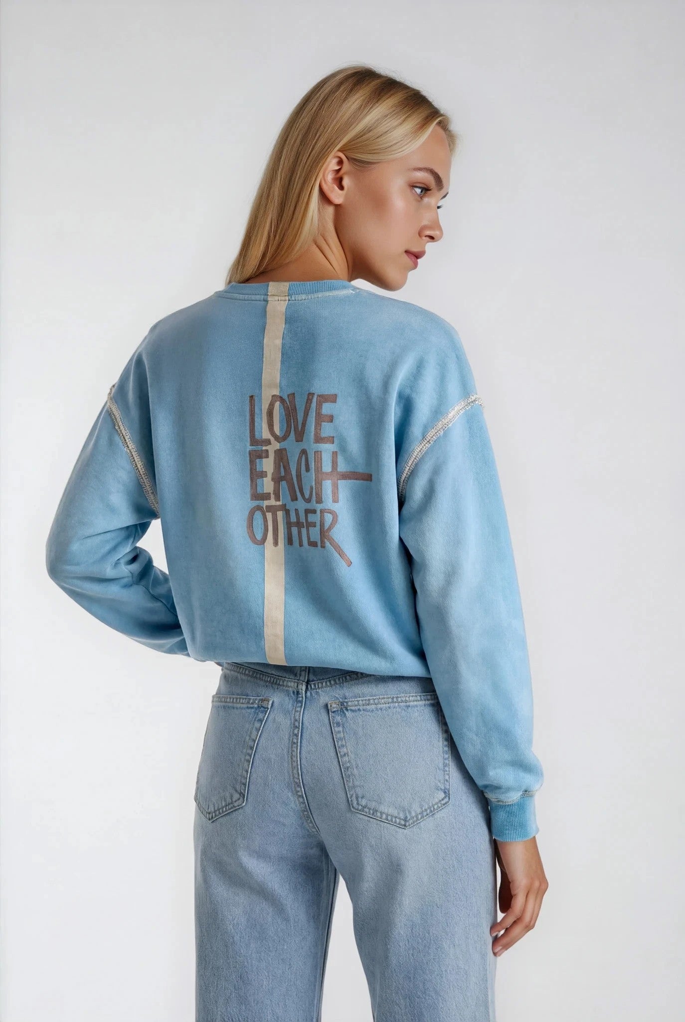 Woman wearing a light blue sweatshirt with 'LOVE EACH OTHER' text on the back, standing against a plain background.